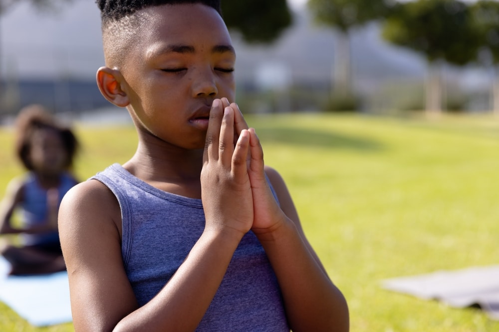 Happy,African,American,Schoolboy,Doing,Yoga,And,Meditating,On,Field